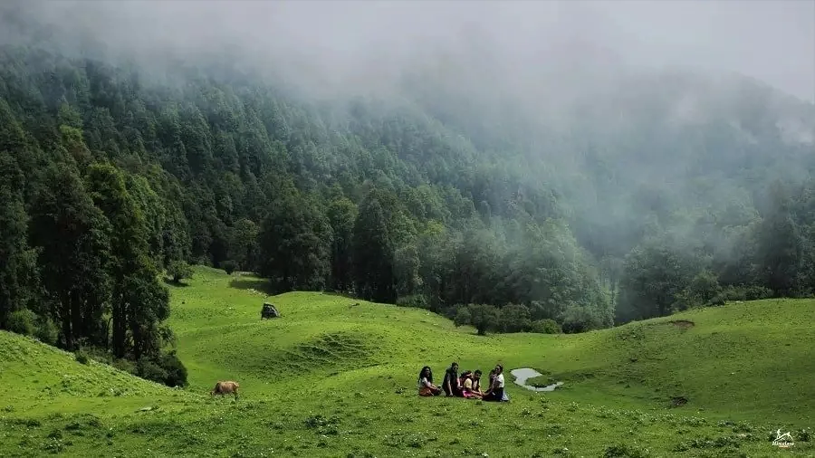 Mist over the forest and a green meadow on the Dayara Bugyal trail, with trekkers resting on the grass in monsoon weather