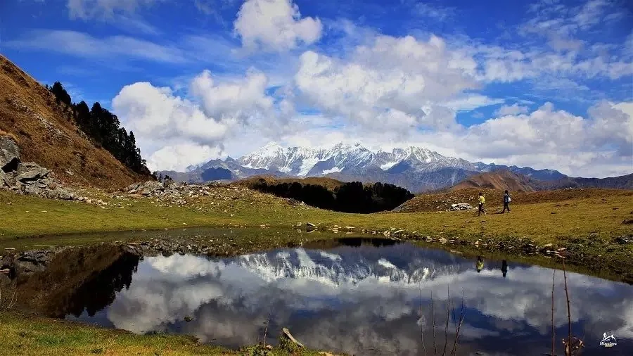 Two trekkers walking past a small pond that reflects snow peaks at Dayara Bugyal under a clear autumn sky