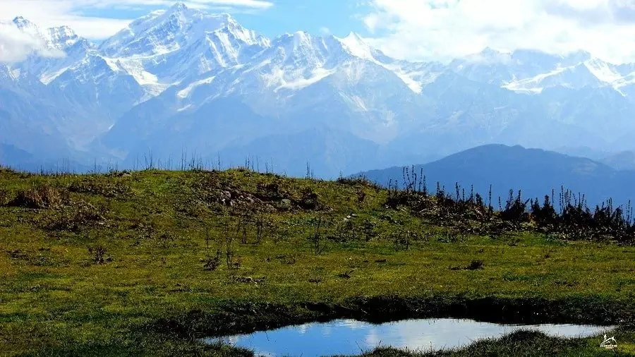 Green grass and a small pond on Dayara Bugyal meadow, with snow-capped Himalayan peaks behind on a clear day