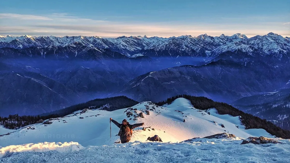 Trekker on the snowy Kedarkantha summit at sunrise, with a long line of Himalayan peaks on the horizon