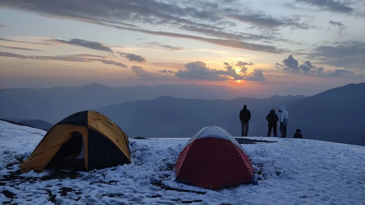 Two tents on snow at a Brahmatal campsite at sunset, with trekkers watching the light fade as the temperature drops