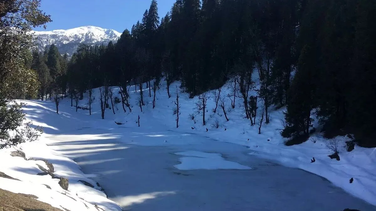 Frozen Juda Ka Talab lake on the Kedarkantha trek, surrounded by snow and tall pine forest under a blue winter sky