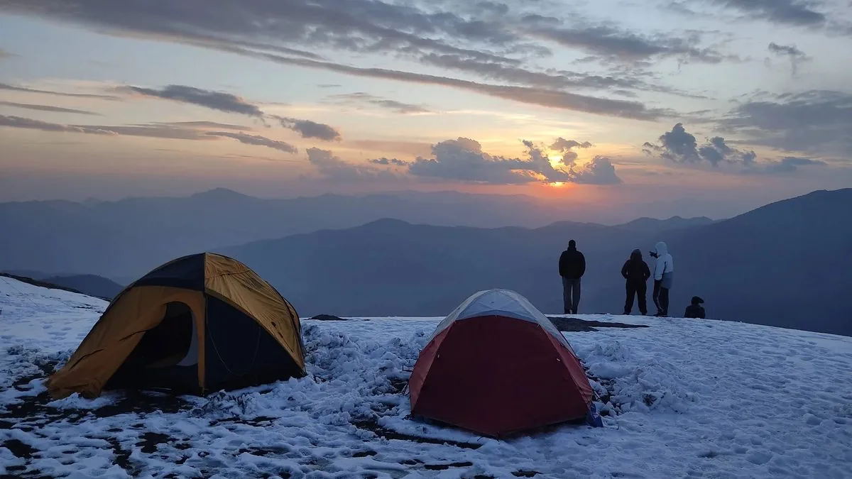 Two trekking tents pitched on snow at a Brahmatal campsite at sunset, with trekkers watching the sky over the hills