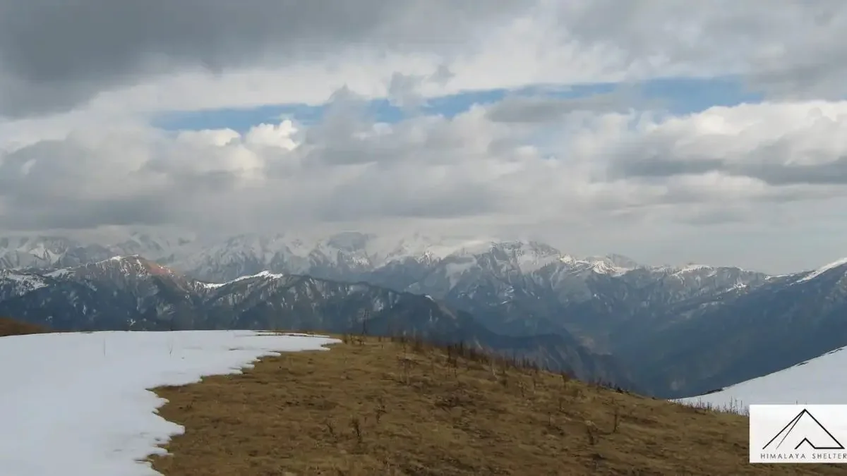 Edge of the Dayara Bugyal meadow with melting snow and brown grass, facing a long range of snow-capped peaks under clouds