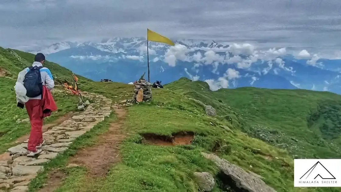 Trekker walking on a stone trail across a green ridge on the Panch Kedar trek, with prayer flags and snow peaks behind