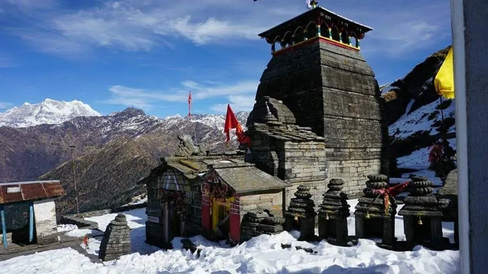 Tungnath stone temple and small shrines with snow on the ground and Himalayan peaks on the left