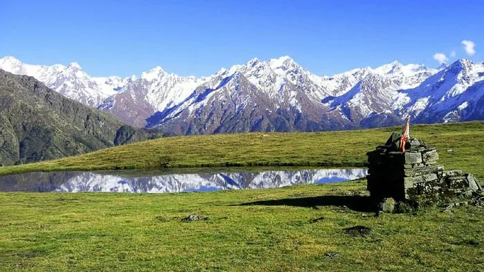 Small stone shrine beside a still pond on a green meadow above Madhyamaheshwar, with a long line of snow peaks behind